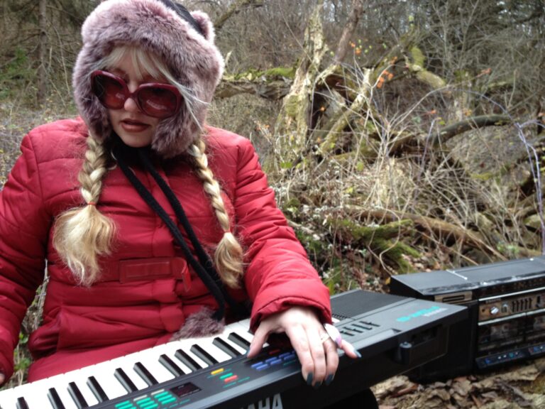 Woman in red coat and fur hat plays keyboard in a wooded area.