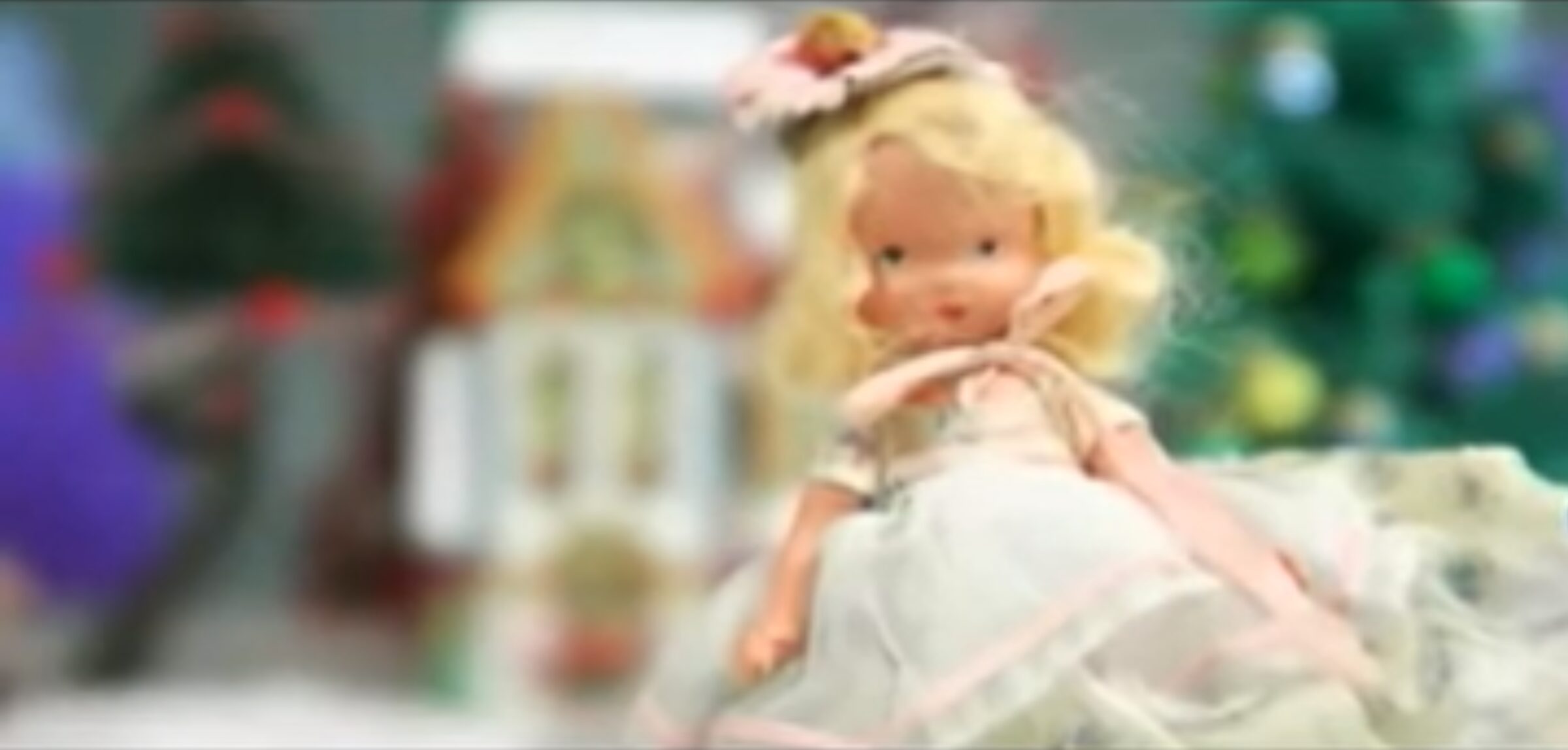 Vintage doll with blonde hair in a pink dress, with a blurred Christmas tree and gingerbread house in the background.