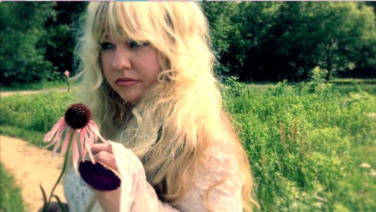 Woman with blonde hair holding a pink coneflower in a grassy field.