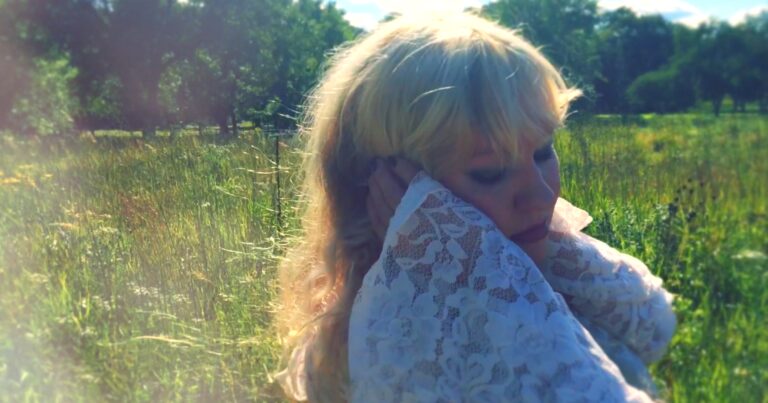 Blonde woman in white lace embraces herself in a sunlit grassy field.