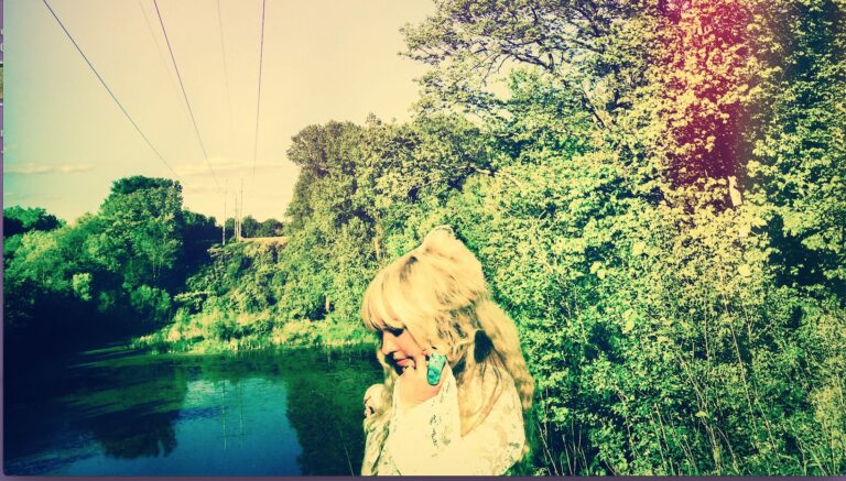 Blonde woman in white lace top with turquoise ring by a river and trees.
