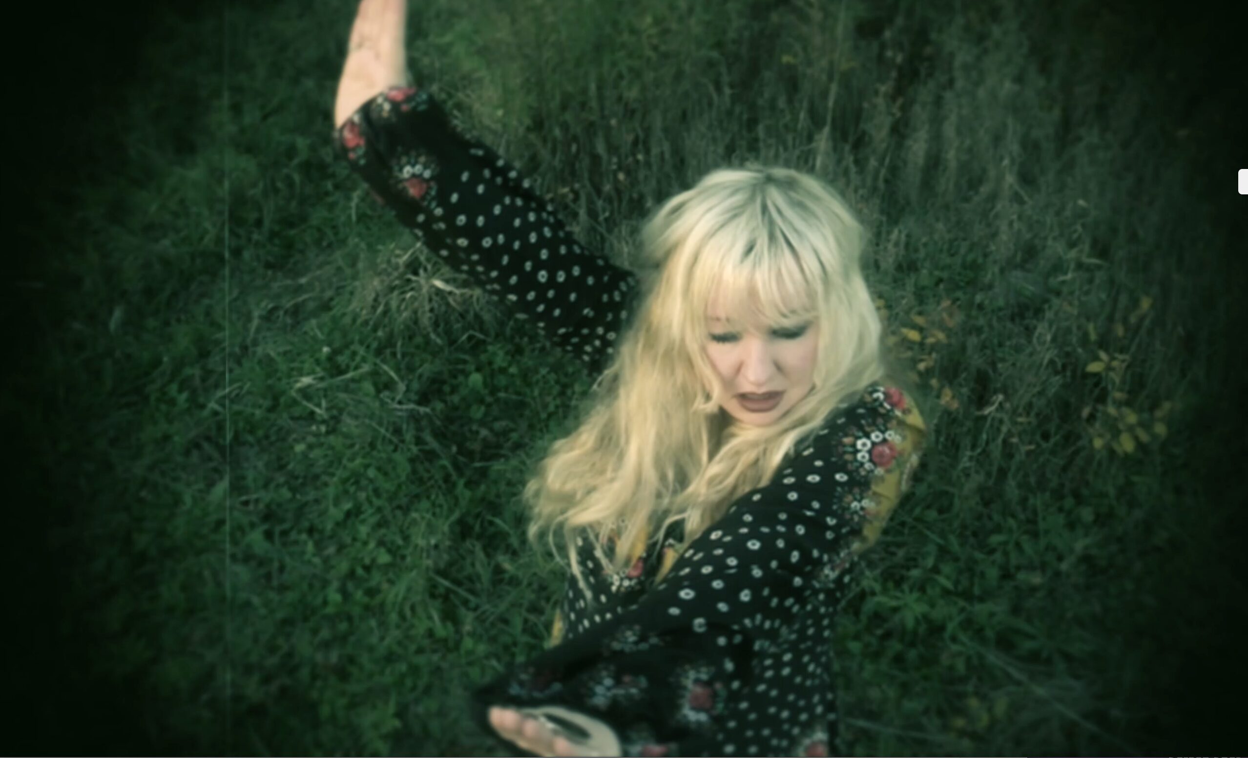 Blonde woman in a black floral dress lying in tall green grass.