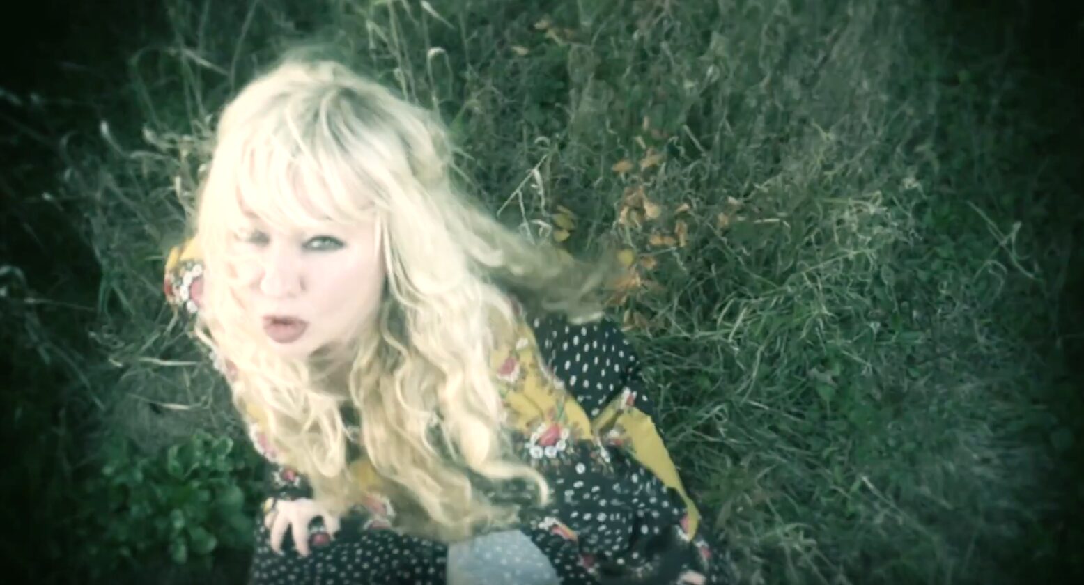 Blonde woman with curly hair in a patterned dress, looking up from tall grass.