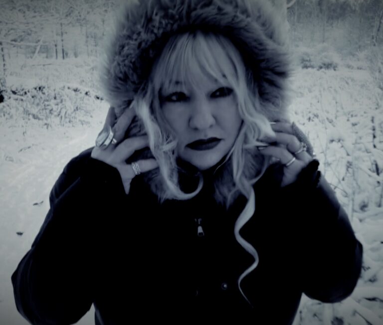Woman with blonde hair and dark lipstick in a fur-hooded coat in a snowy landscape.