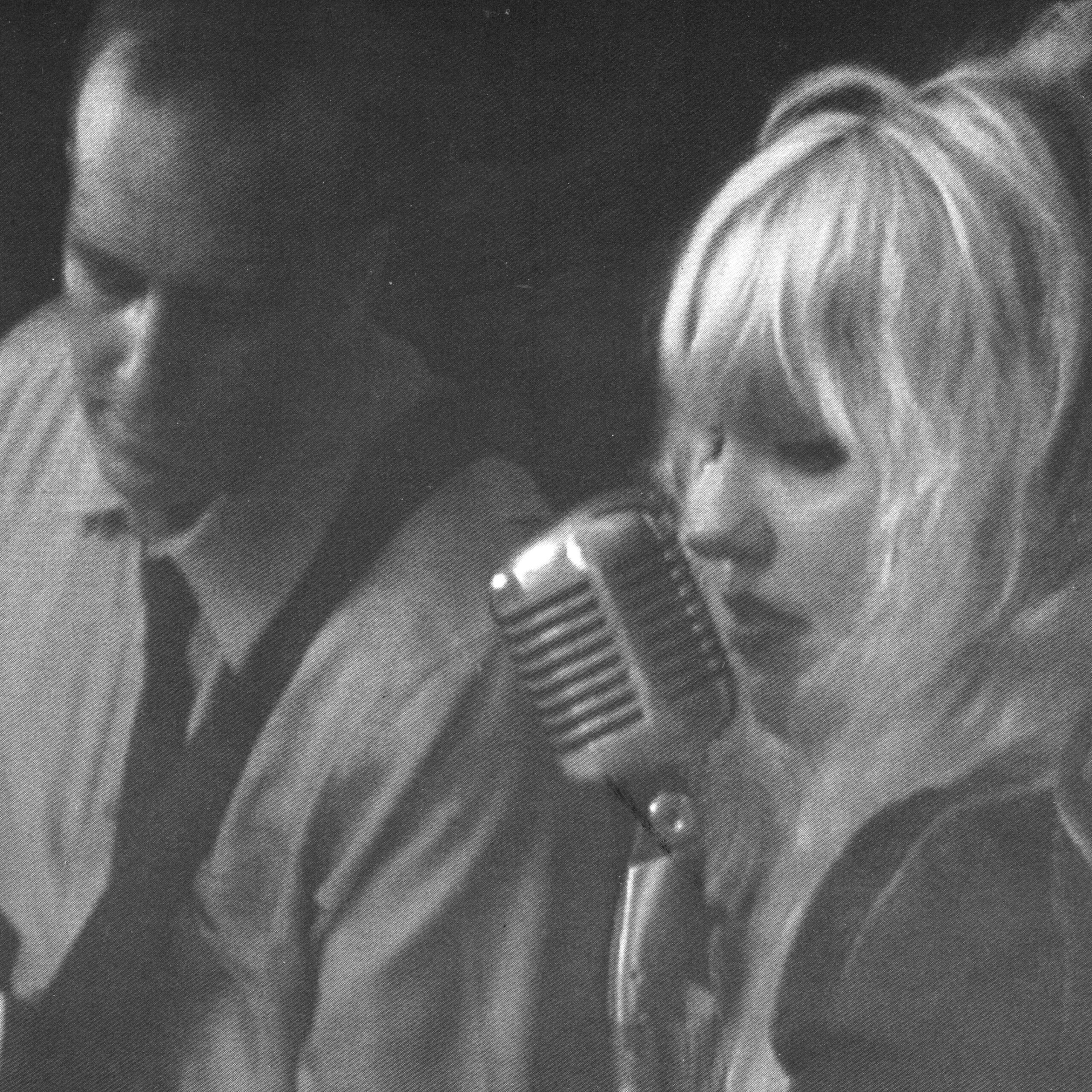 Woman with blonde hair singing into a vintage microphone, man in background.