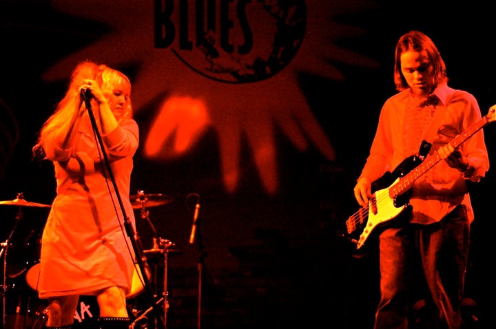 Singer with blonde hair holding a microphone and a man playing a bass guitar on stage.