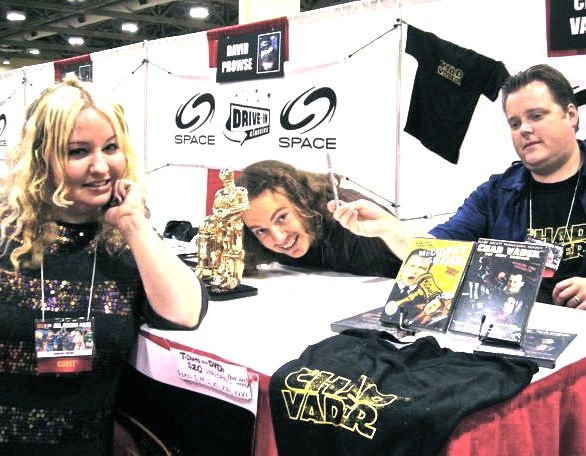 Three people at a table with Darth Vader merchandise, including DVDs and t-shirts.